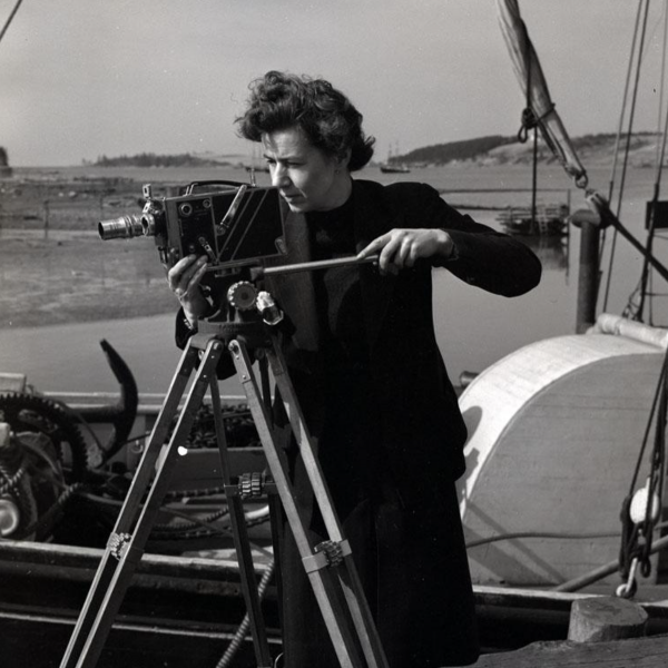 A black and white archival photograph of a woman operating a film camera on a tripod standing in front of a boat.