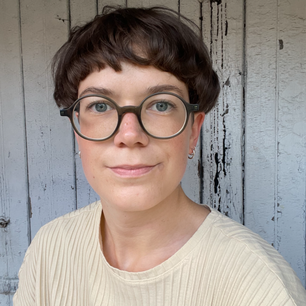 A photograph of a woman with round glasses and short dark brown hair who is wearing a beige t-shirt and who is standing in front of a wooden wall with peeling white paint.