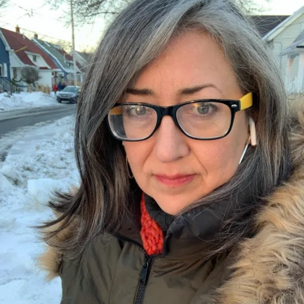 A photograph a woman with black and yellow glasses who is wearing a green and brown parka and standing outside on a residential street during the daytime in the winter.