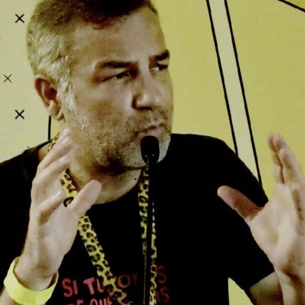 A photograph of a man with short hair speaking in front of a table microphone. The man is gesticulating with both hands and the photo has been taken while he is speaking. 