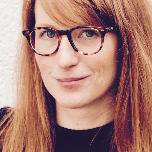 A portrait photograph of a smiling woman with auburn hair, brown horn-rimmed glasses, and a black shirt.