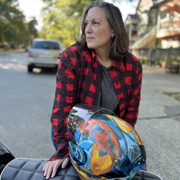 A colour photograph of a woman in a red and black flannel shirt standing in front of a motorcycle with a colourful orange and blue helmet resting on the seat.