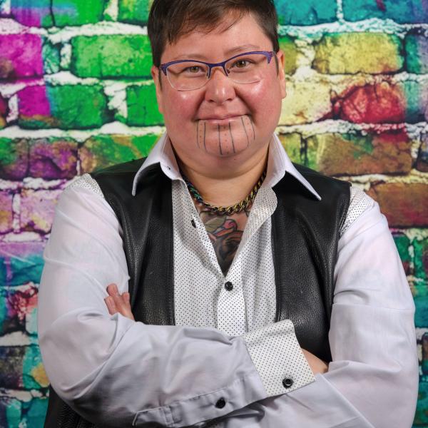 A portrait photo of a man in a light grey collared shirt and a black leather vest standing in front of a colourfully painted brick wall.