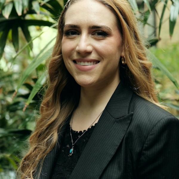 A portrait photograph of a woman with long light brown hair who is wearing a black blazer and is standing in front of a bunch of tall, tropical plants.