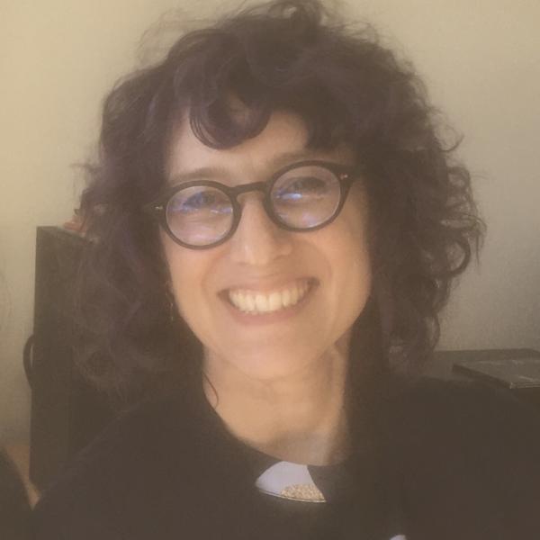 A colour photograph of a smiling woman with black glasses, black curly hair, and dark clothing.