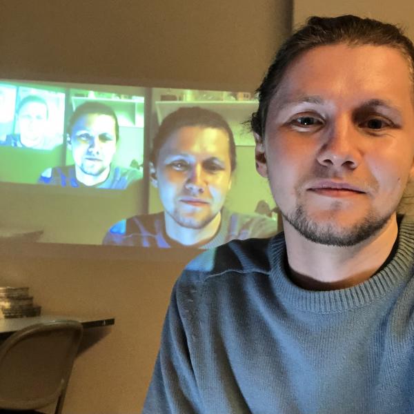 A photo of a man with tied back brown hair and a goatee sitting in front a video projection of himself.