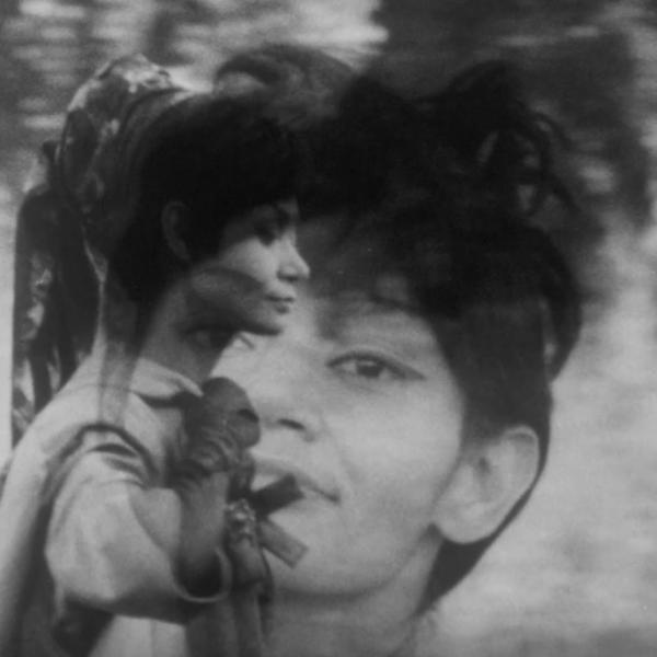 A black and white film still of a woman's face overlaid onto of a image of a woman walking in a forest during the day toward the right of the frame.