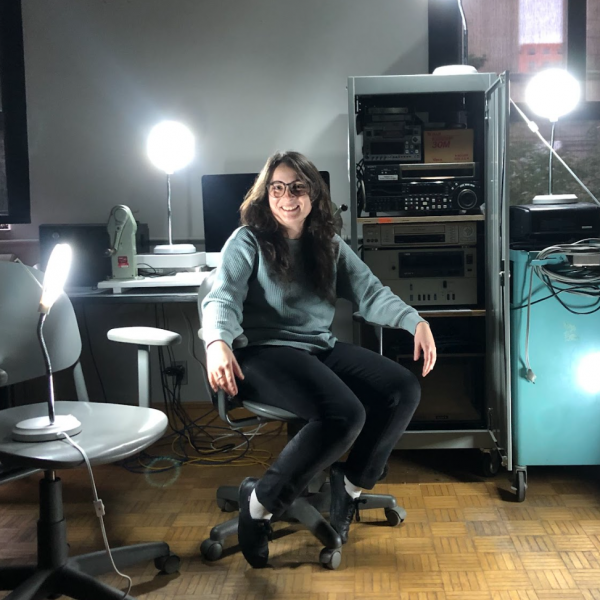 A colour photograph of a woman sitting in a office who has long brown hair, a light grey sweater, and dark paints and is sitting in an office chair. There are lots of audiovisual equipment and lamps arranged around them.