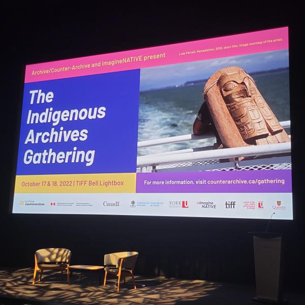 A photo of a dimly lit theatre stage with two chairs and a small table and a large colour projection on the wall behind with the text "The Indigenous Archives Gathering" displayed.