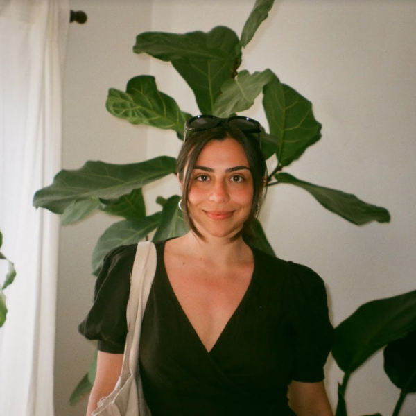 A photograph of a young woman with dark brown hear who is wearing black top, holding a beige tote bag, and standing in a white room with lots of tall plants.