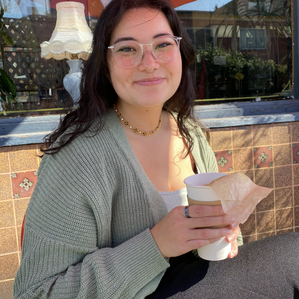 A photograph of a young woman with long black hair, clear glasses, and grey clothing who is sitting outside with a coffee in front of a window on a sunny day.