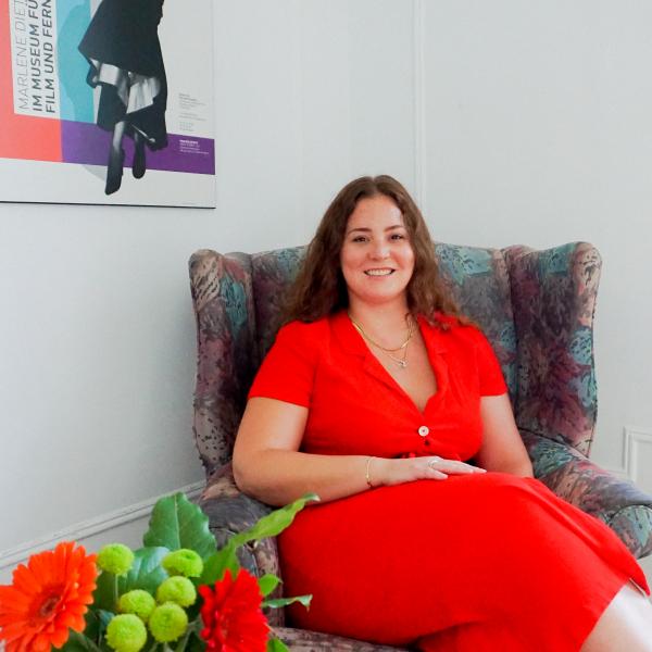 A colour photograph of a young woman who is wearing a bright red dress and is sitting on a grey upholstered chair with flowers on a table in front of them and a colourful poster on the wall to their right.