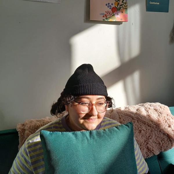 A photograph of a young woman sitting on a blue-green couch. She is wearing wire-frame glasses, a black toque and a striped shirt. The white wall behind her has art hung on it and light coming in from a nearby window.