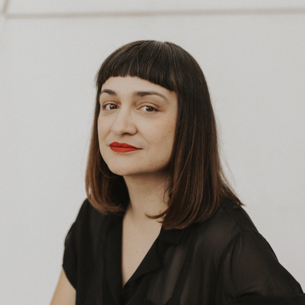 A photograph of a woman with long dark brown hair, red lipstick, who is wearing a black blouse, and standing in front of a plain white wall.