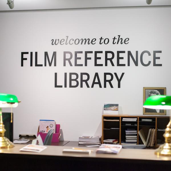 A colour photograph of a desk in front of a white wall. The wall has the text "welcome to the FILM REFERENCE LIBRARY" written in 3D black text.