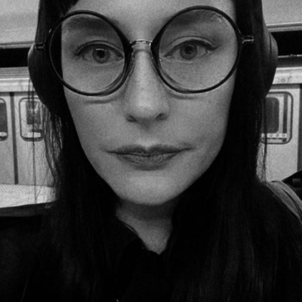 A black and white portait photograph of a woman with large round glasses and dark hair standing in an underground subway station.