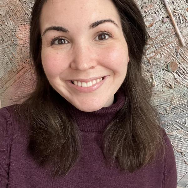 A self-portrait photo of a young woman with shoulder-length dark brown hair who is standing in front of a beige, roughly-textured wall. She is wearing a purple turtleneck and is smiling.