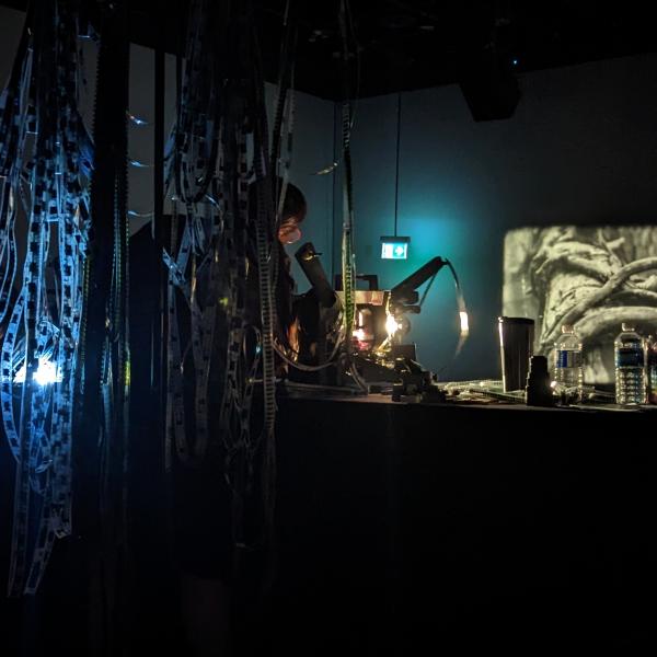 A photograph taken of a woman operating several analog film projectors in a dark space. There is a black and white projection in front of her and long strands of film reel hanging from behind her.