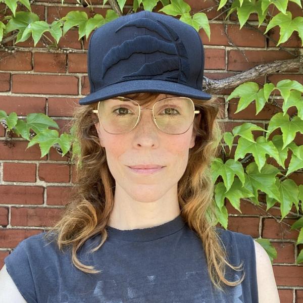 A sqaure selfie portrait photograph of a woman with long light brown hair, wireframe glasses, and a black baseball cap. The woman is standing in front of red-brick wall with ivy vines growing on it. 