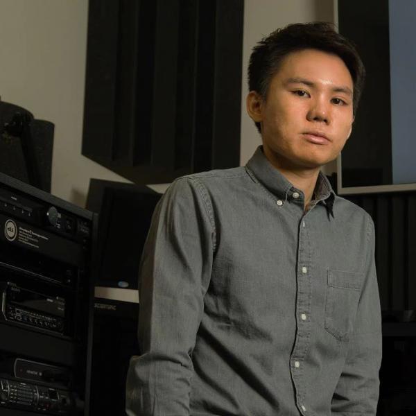 A photograph of a person with short dark hair and a grey collared shirt with white buttons. They are standing in a dim room with AV equipment behind them.