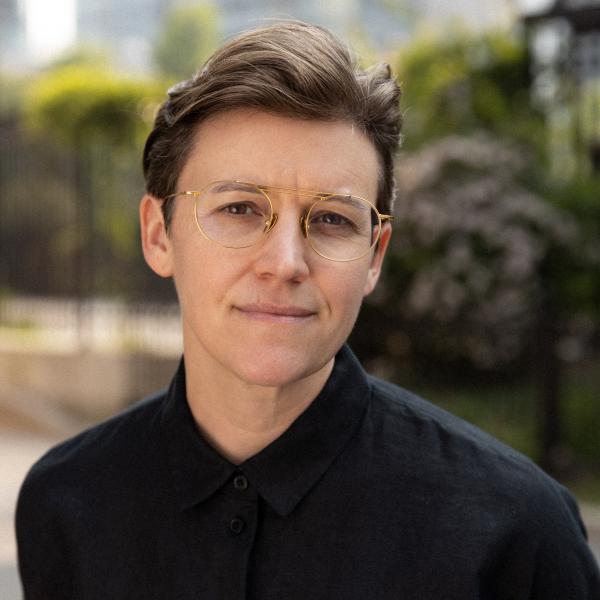 A photo of a person with glasses, shirt dark brown hair, and wireframe glasses standing outside on a road in front of some trees and pushes during the daytime.