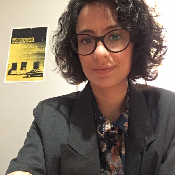 A photo of a woman who is sitting in a well-light interior space with white walls. She has dark curly hair, large black glasses, and is wearing a dark grey blazer and a patterned shirt.