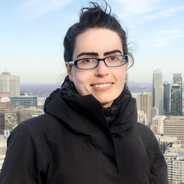 A portrait photo of a woman with black hair and glasses standing in front of a city skyline on a winter day.