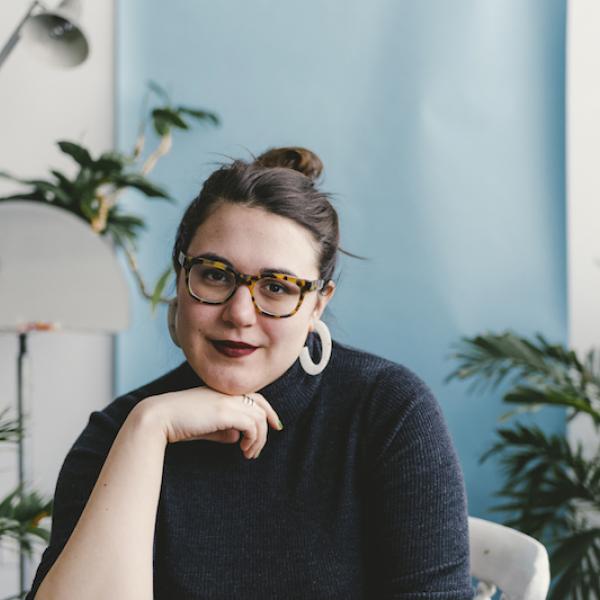 A portrait photograph of a young woman sitting in a white interior space. She is has glasses, dark hair, a black shirt, and is resting her chin on her right hand.