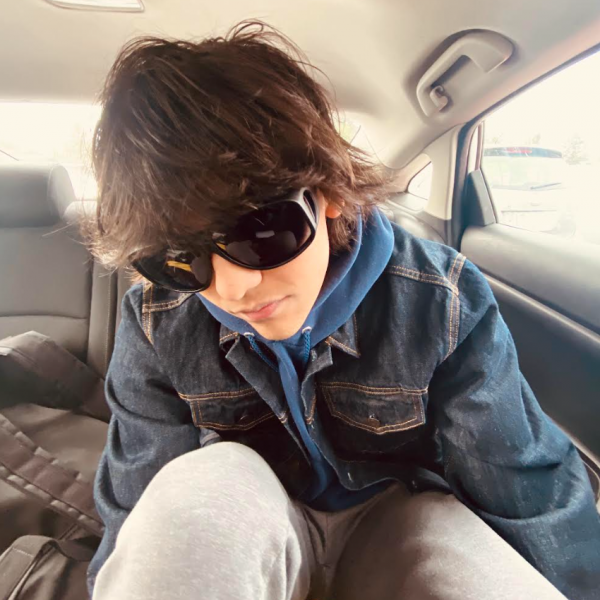 A photo of a young man sitting in the back seat of a car. He is wearing large dark sun glasses, had shaggy dark brown hair, and is wearing a blue denim jacket.