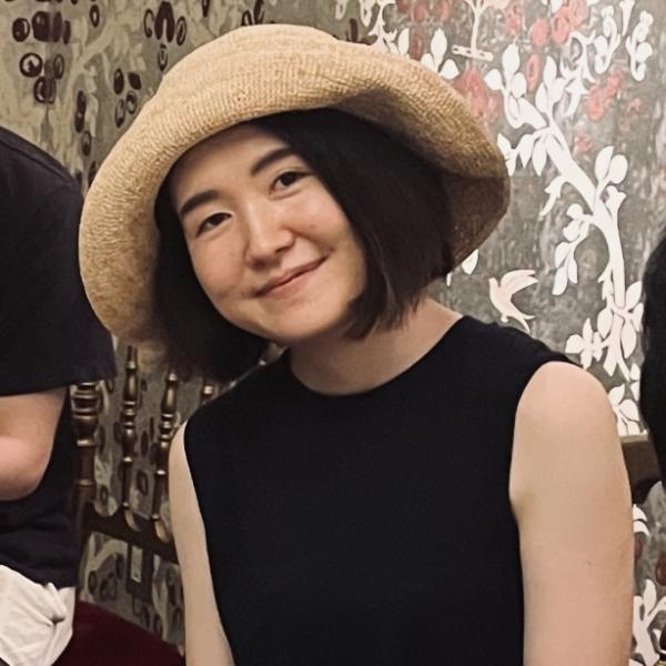 A photograph of young smiling woman who has dark, medium-length straight hair and is wearing a beige sun hat and a black sleeveless top. She is sitting in a room with other people and a floral tapestry in the background.