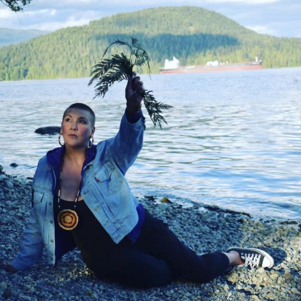 A photograph of a woman sitting on a rocky beach in front of a large lake with a forested small mountain in the background. The woman has very short dark hair and is wearing a denim jacket and holding a bundle of plants in the air.
