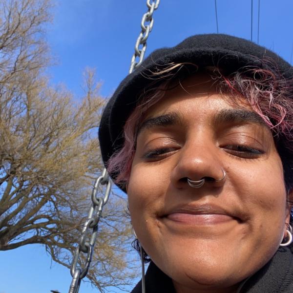 A selfie photograph of a smiling person sitting on a swing set on a clear sunny day.
