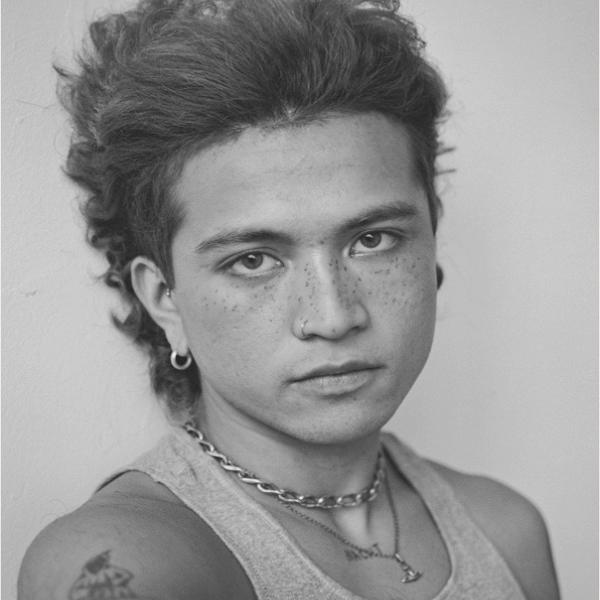 A black and white portrait photograph of a young person with wavy hair, a tanktop, a few necklaces, and earrings. They are standing in front of a white wall and are no smiling.