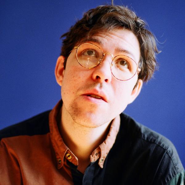 A colour photograph of a person with a multicolour patchwork patterned collared shirt who has short brown hair and wireframe glasses. They are standing in front of a plain blue wall and looking up away from the camera.