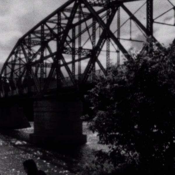 A dark black and white film stilll of an old metal bridge on a cloudy day.