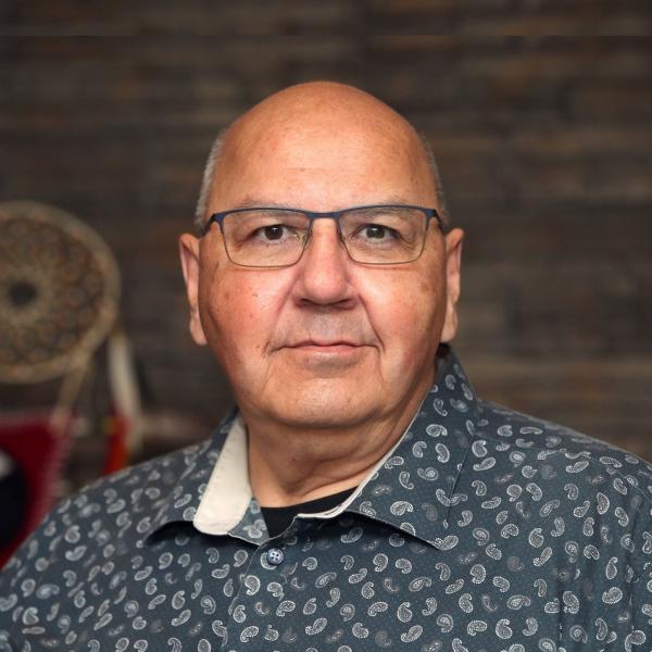 A portrait photograph of a man shot against a dark, brown background. He looks into the camera and wears a blue patterned shirt and wire-rim glasses. The subject is bald and has dark eyes. The photograph is professional and well lit. 