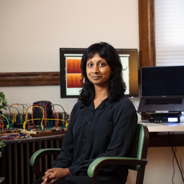 A striking, coloured photograph of a woman sitting at a desk. She looks at the camera with her hands clasped in her lap. Behind her, the desk is covered in colourful electronics. The woman has dark hair with bangs parted in the middle. She is wearing a dark button-down shirt. She sits in a green chair and various plants adorn her desk. 