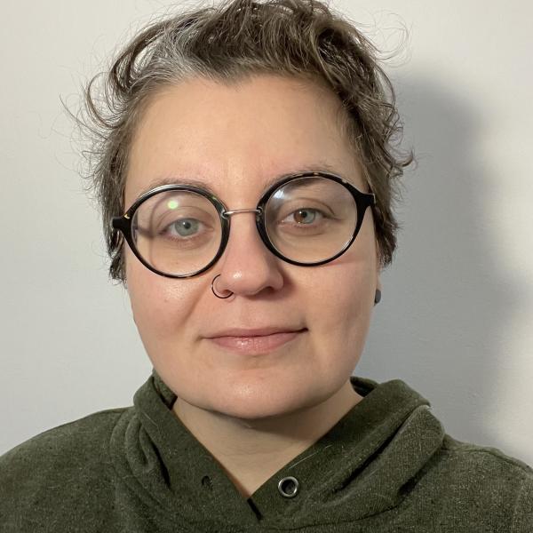 A portrait photograph of a person with black, rounded glasses, smiling slightly at the camera, mouth closed. The image is a close-up of the subject's face and shoulders. They wear a hooded sweatshirt and have short, grey, curly hair. Their eyes are blue with a brown patch in the iris of their left eye. 