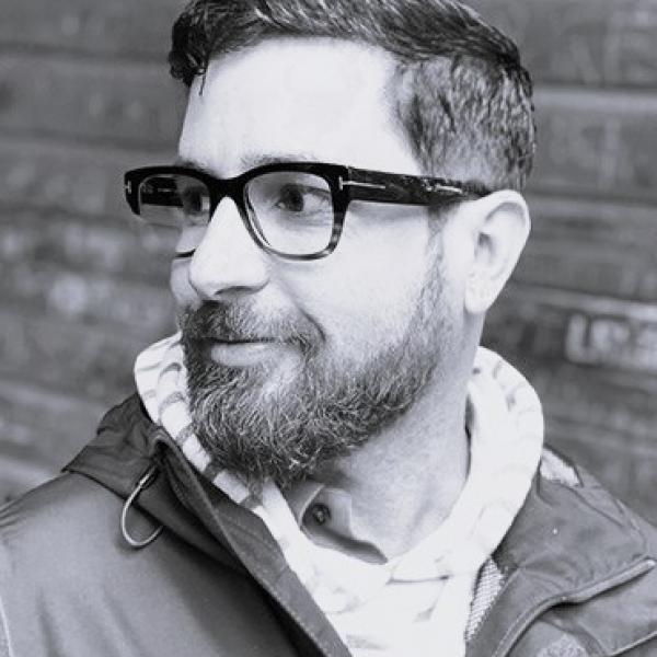 A black and white portrait photograph of a man shot against a brick background. He wears a hooded sweatshirt underneath a windbreaker jacket. He is shot in partial profile and has short hair and a beard. He wears thick-framed glasses and smiles slightly. 