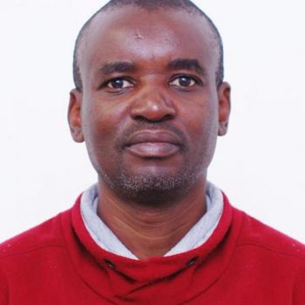 A passport-style photograph of a man with short dark hair and a short goatee staring straight ahead into the camera. He is wearing a red sweatshirt and white collared shirt and is standing in front of a white background.