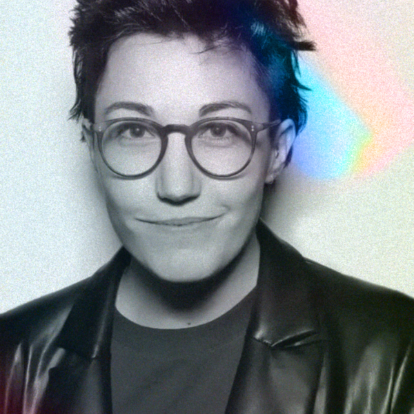 A black and white portrait image of a person in front of a white background. There is a coloured, rainbow effect that highlights the left side of the person's face. They have short brown hair and are wearing a leather blazer over a T-shirt. They are smiling while looking directly into the camera. 