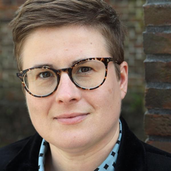 A close-up portrait image of a person staring into the camera. They wear tortoiseshell horn-rimmed glasses and have short, light brown hair styled to one side. They have dark brown eyes and wear a patterned blue shirt underneath a black blazer. They are shot against a red brick background. 