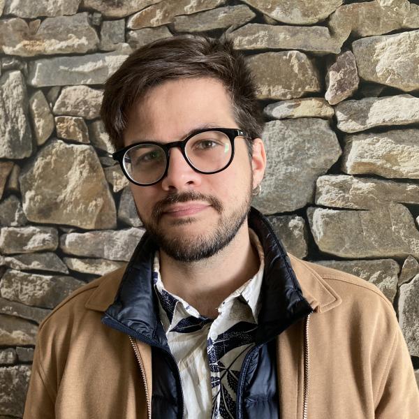 A man photographed against a grey stone wall. He has short, dark hair and a short beard. He wears black glasses with a thicker frame, along with a patterned button down shirt, a black jacket, and a brown jacket layered over one another. He has a silver hoop earring in one ear and looks straight into the camera. 