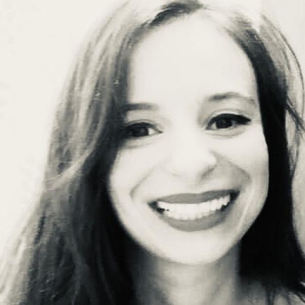 A high-contrast black and white photograph of a woman's face. She looks into the camera with a big smile on her face and has long hair, parted to one side. She is pictured against a white background. 