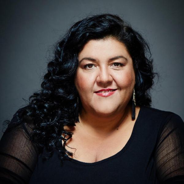 A striking professional photographic portrait of a woman against a moody grey background. She smiles slightly and has long, dark, curly hair. She wears a black top with transparent sleeves and ornate earrings.  