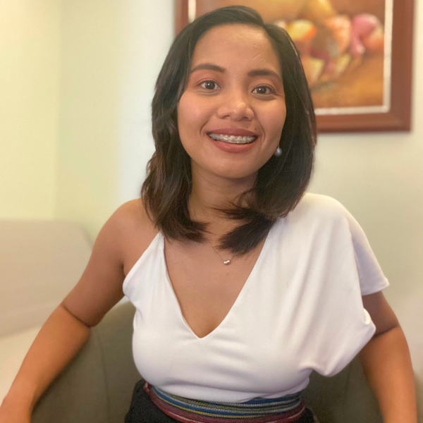 A square portait photo of a smiling woman with shoulder-length dark hair who is wearing an asymmetrical white top with one arm that is sleeveless. They are sitting in a room with a framed still life behind them.