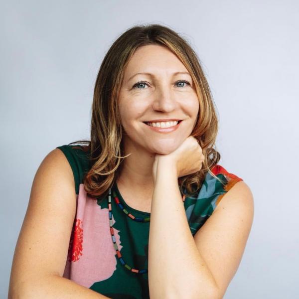 A professional potrtrait of a woman looking into the camera, against a solid grey background. She smiles brightly and rests her chin on her hand. She has dark blonde hair and wears a green top covered in large, colourful flowers with a necklace of multi-coloured beads. Her eyes are blue. 