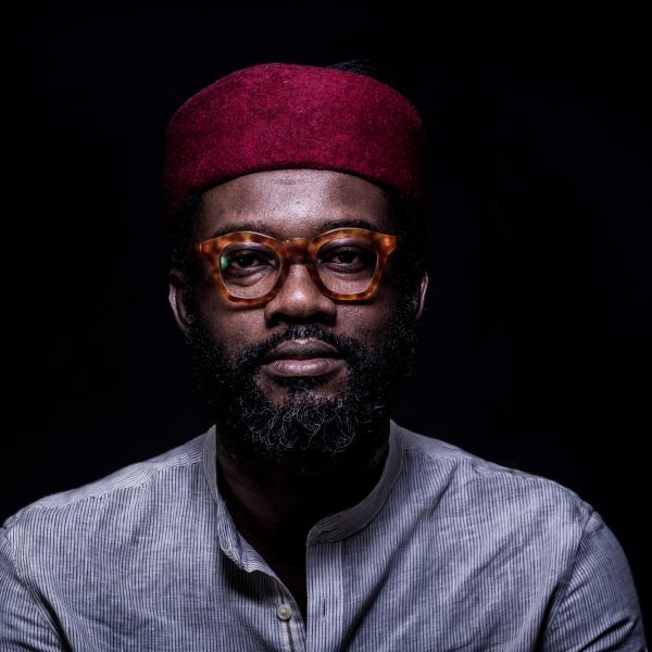 A portrait photograph of a man in horn-rimmed, tortoise shell glasses. They are wearing a red hat and a cotton button down shirt with thin stripes. The subject is lit from a single light source against a black background. 