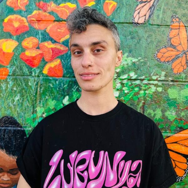 A portrait photo of a person in a black graphic T-shirt against a colourful background. The subject smiles slightly and looks at the camera.They stand in front of a painted mural that depicts a colourful display of orange flowers and monarch butterflies. 