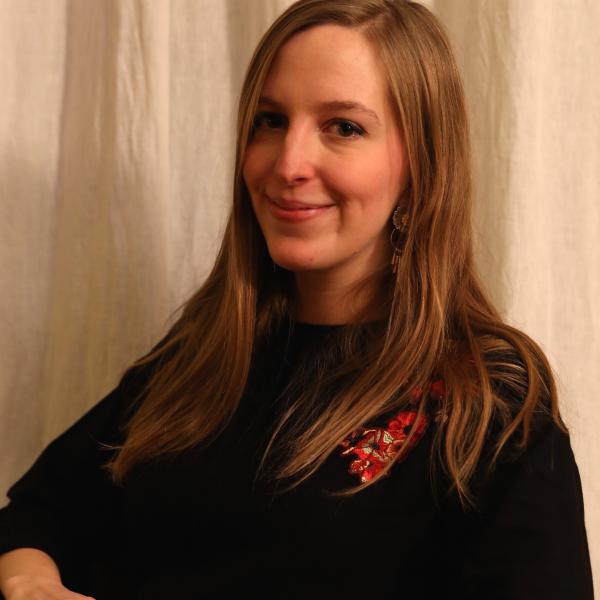  A portrait photo of a young woman with long straight light brown hair who is wearing a black sweater and sitting in front of some light brown curtains. She is smiling and has her hands clasped.